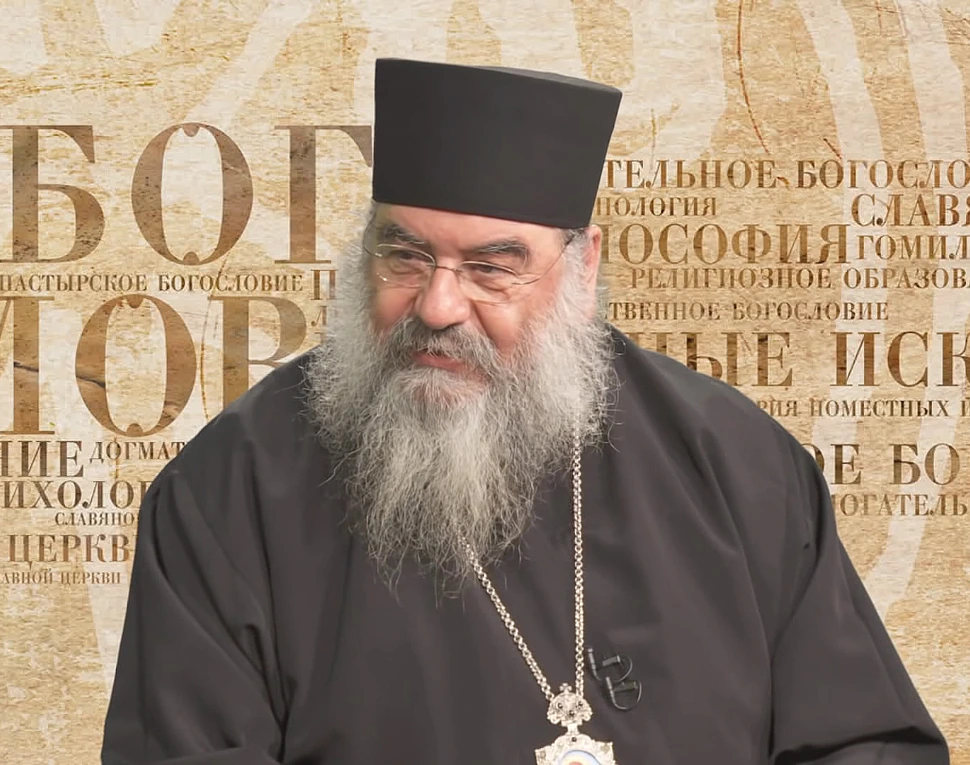 Orthodox cleric Athanassios Kyros Filippou in black robes and hat, speaking during an interview against a patterned back